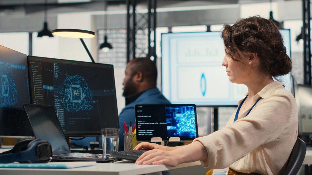 A woman working at a desk with multiple monitors displaying AI graphics and code in a modern office, with a colleague working in the background.